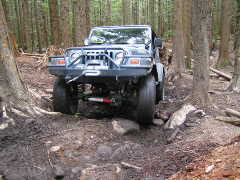 1990 Ford Bronco - A Jeep Wrangler navigates a challenging off-road course through a dense forest with muddy terrain and large rocks.