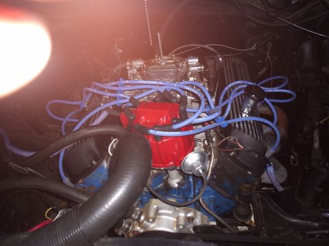1985 Ford Bronco - Blue-block V8 photographed under artificial light with no air cleaner, showing an aluminum four-barrel carburetor, a red HEI-style distribu…