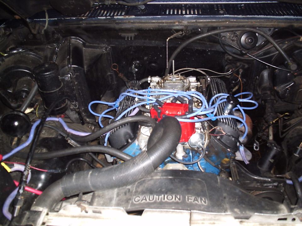 Blue V8 with finned valve covers, bare four-barrel carb, blue plug wires and CAUTION FAN shroud