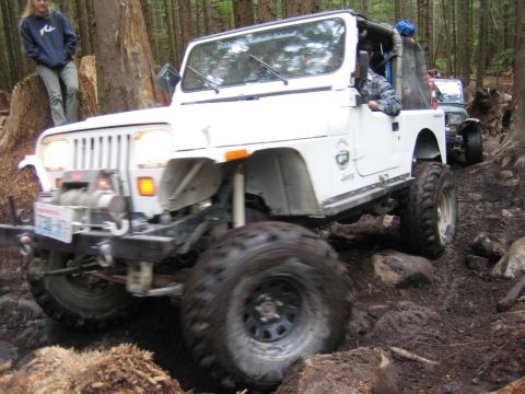 1990 Ford Bronco - A white Jeep navigates a challenging, muddy off-road course with trees all around.