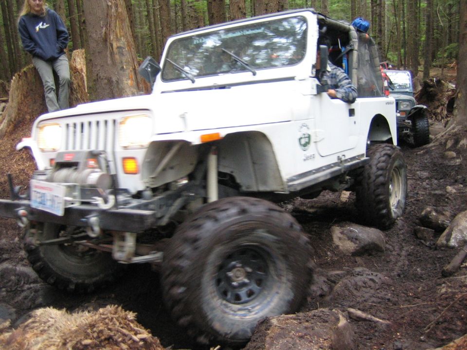 White Jeep Wrangler driving on a muddy, wooded trail. Another Jeep is visible behind it