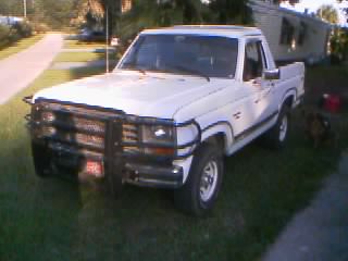 1986 Ford Bronco - Low-resolution three-quarter front view of a white 1980s Ford regular-cab pickup with a black tube brush guard and driving lights, chrome m…