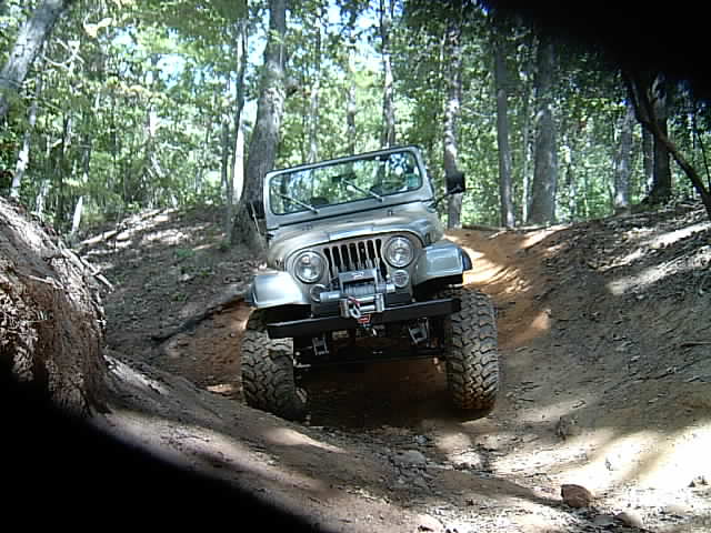 Silver Jeep CJ with front winch crawling a wooded trail rut, front tire stuffed