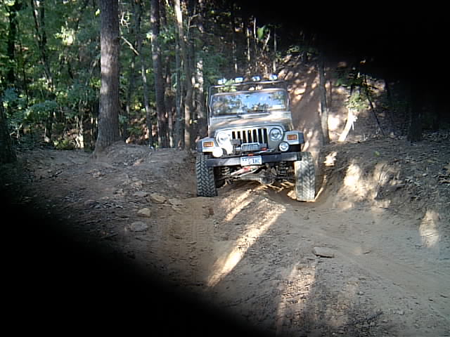 Silver Jeep Wrangler TJ with light bar climbing a sunlit dirt rut in the woods