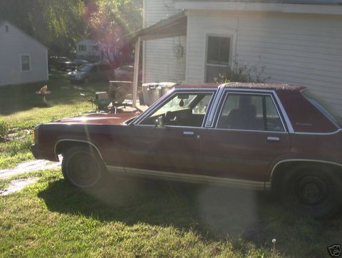 1991 Ford LTD Crown Victoria - A dark red four-door Ford LTD Crown Victoria sedan photographed from the driver's side in a grassy yard beside a white house, with sun flar…