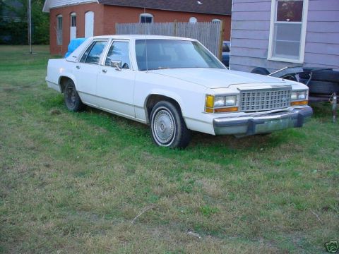 1991 Ford LTD Crown Victoria - A white box-body Ford LTD Crown Victoria four-door sedan sitting in a grassy yard, shot from the 3/4 front on the passenger side, with a re…