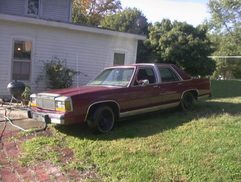 1991 Ford LTD Crown Victoria - A dark red Ford LTD Crown Victoria four-door sedan parked on grass beside a white clapboard house, seen from the 3/4 front on the driver's…