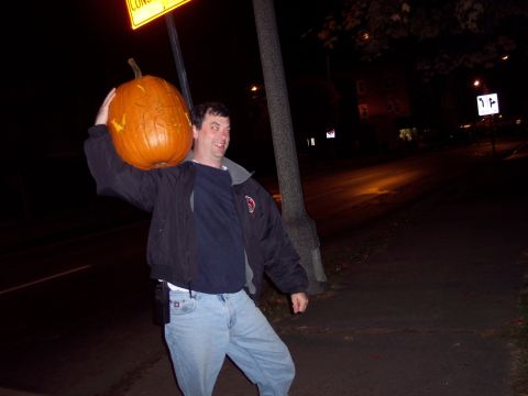 2001 Ford Crown Victoria - A man carries a large carved pumpkin over his shoulder at night, with streetlights and a sign visible.