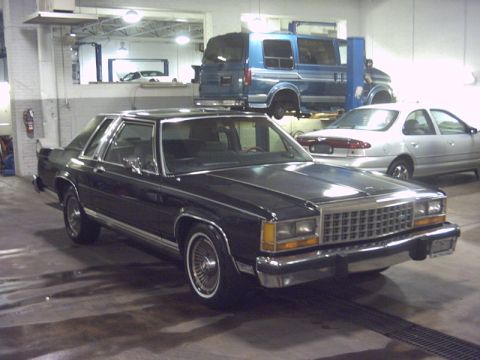 1991 Ford LTD Crown Victoria - A dark blue 1980s Ford Crown Victoria is parked in a garage, with a blue van on a lift in the background.