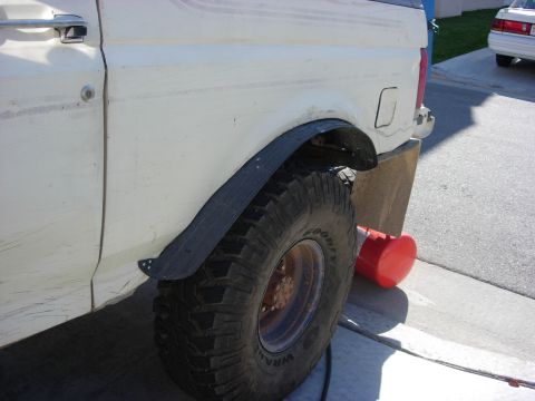1987 Ford Bronco - Side view of a 1987 Ford Bronco's rear fender and tire, showing custom fender flares. A second vehicle is parked nearby.