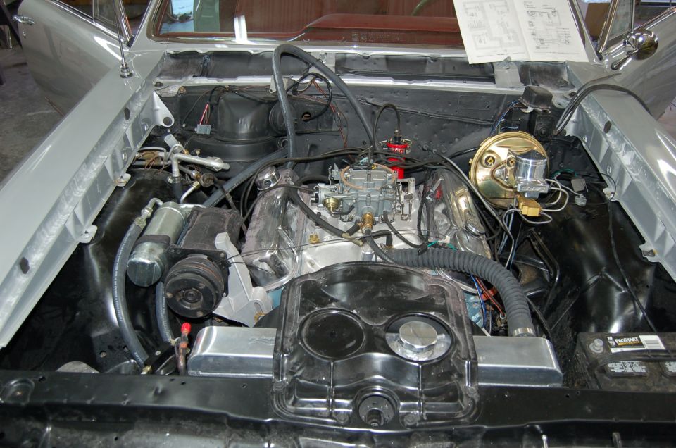 engine bay with V8 engine, carburetor, and air conditioning unit. Some papers are on the dashboard