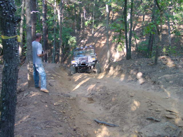 Man in gray shirt watching a Jeep Wrangler climb a rutted dirt hill in the woods