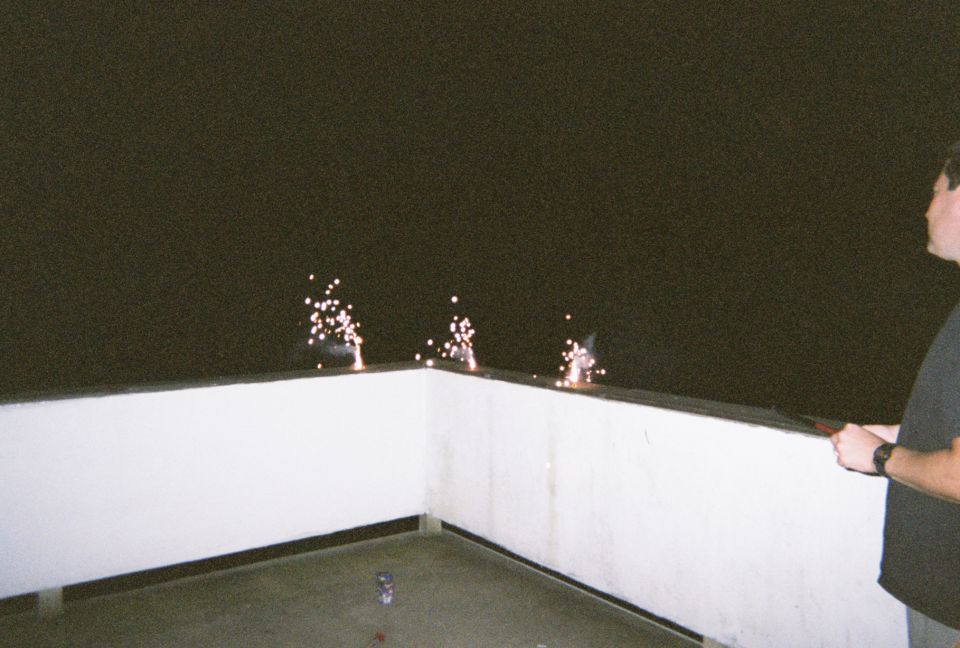 Person holding a sparkler near fireworks on a white railing at night