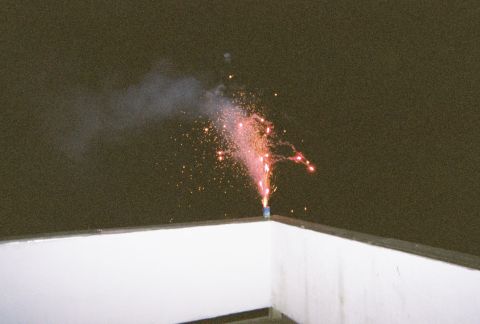 1990 Ford Bronco - A firework erupts with sparks and smoke against a dark sky, viewed from a rooftop.