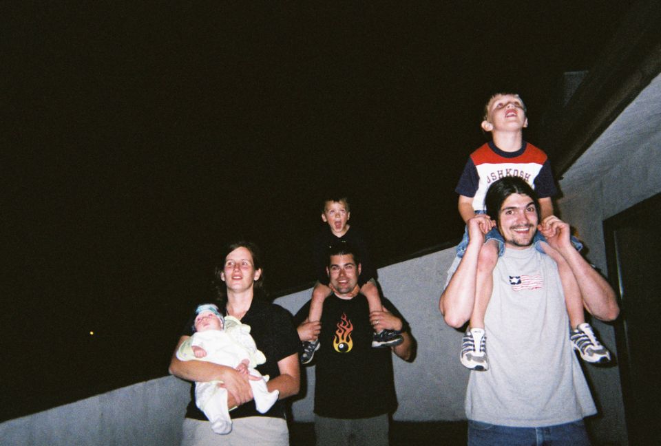 Family members, including adults carrying children on their shoulders, look up at the night sky