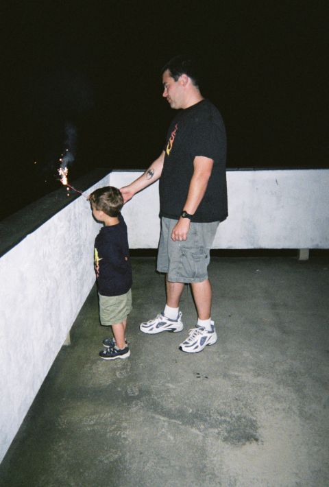1990 Ford Bronco - A man and a child stand on a concrete surface, watching a sparkler ignite against a dark background.