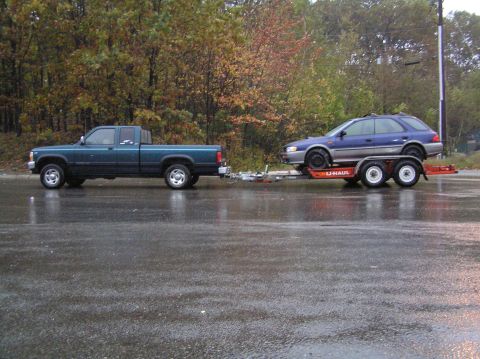 1997 Subaru Impreza - A blue Subaru wagon is being towed on a U-Haul trailer by a pickup truck on a wet road.