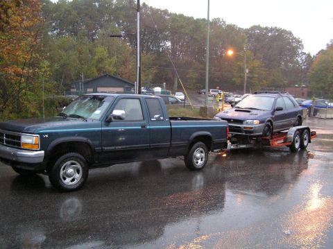 1997 Subaru Impreza - A dark green pickup truck tows a blue Subaru Impreza wagon on a trailer on a wet, overcast day.