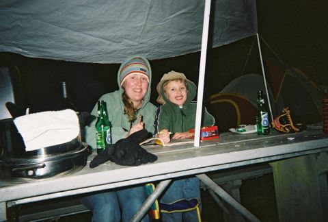 1990 Ford Bronco - A woman and a child sit at a picnic table under a tarp, with camping tents visible behind them.