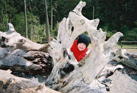 1990 Ford Bronco - A smiling child in a red jacket and black beanie peeks out from a hollow in a large piece of driftwood on a beach.