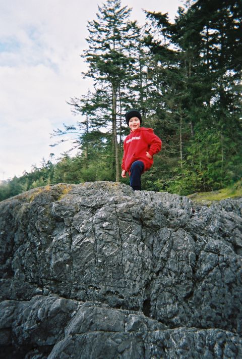 1990 Ford Bronco - A smiling boy in a red hoodie, dark pants and a black knit beanie stands atop a large grey rock outcrop, with tall evergreen trees and an o…