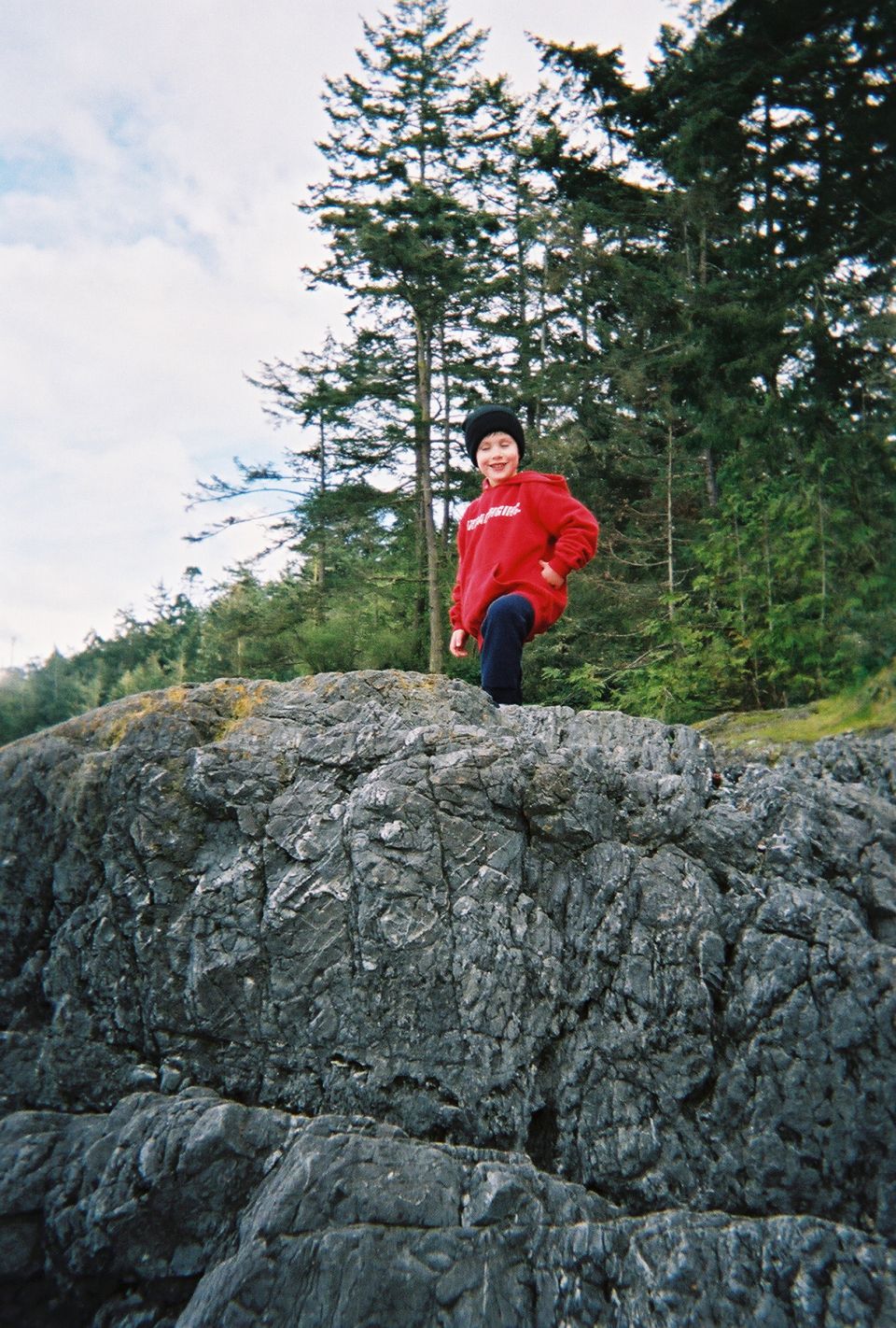 Boy in a red hoodie and black beanie standing on a rock outcrop by evergreens