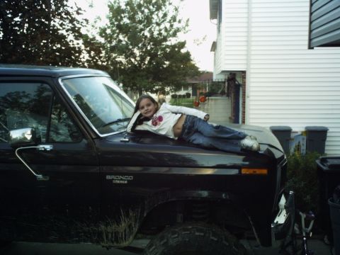 1986 Ford Bronco - A young girl rests on the hood of a black Ford Bronco, with a house and trees in the background.