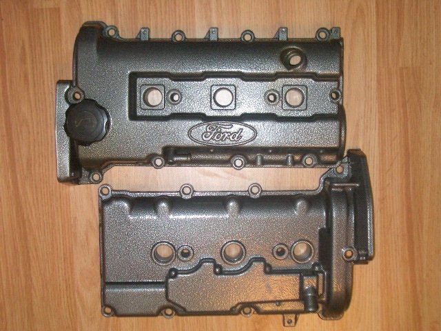 Two gray Ford engine valve covers on a wooden surface. The top cover has a black oil cap and an embossed Ford logo
