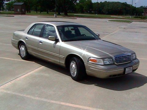 2000 Ford Crown Victoria - A gold/champagne Ford Crown Victoria full-size sedan photographed from the front passenger side (3q-front angle) in an empty concrete parki…