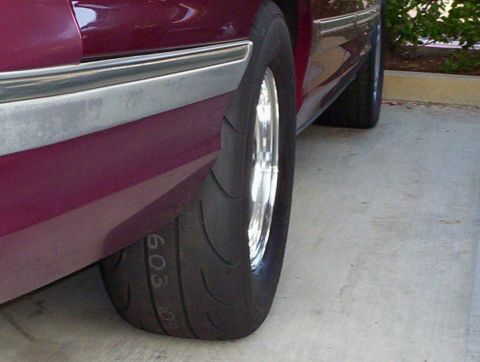 1992 Ford Crown Victoria - Low close-up of the rear wheel of a maroon full-size sedan on a concrete driveway, fitted with a wide soft-compound drag radial on a polish…