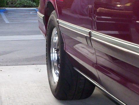 1992 Ford Crown Victoria - Close-up of the front wheel and lower body side of a maroon sedan in a parking lot, showing a narrow tire on a polished aluminum wheel tuck…