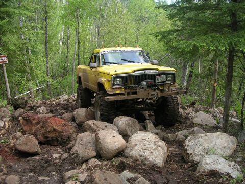 1977 Ford F-150 - A modified yellow Jeep pickup navigates a challenging, boulder-strewn path in a wooded area.