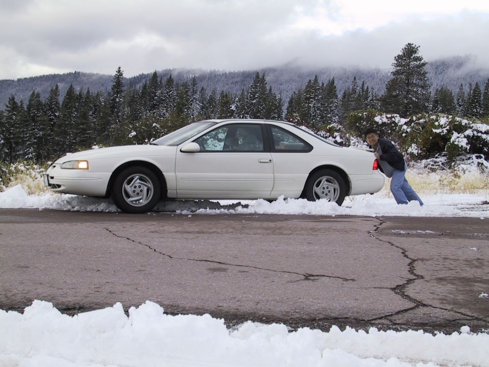 White 1997 Ford Thunderbird stuck in snow, side view. Person pushing