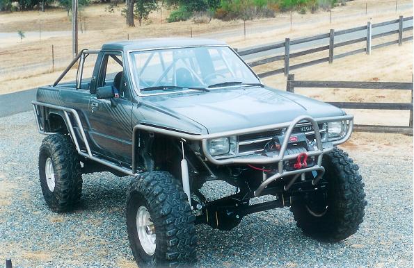 Modified Toyota pickup truck with roll cage, large tires, and custom bumper. Appears to be from the late 1980s