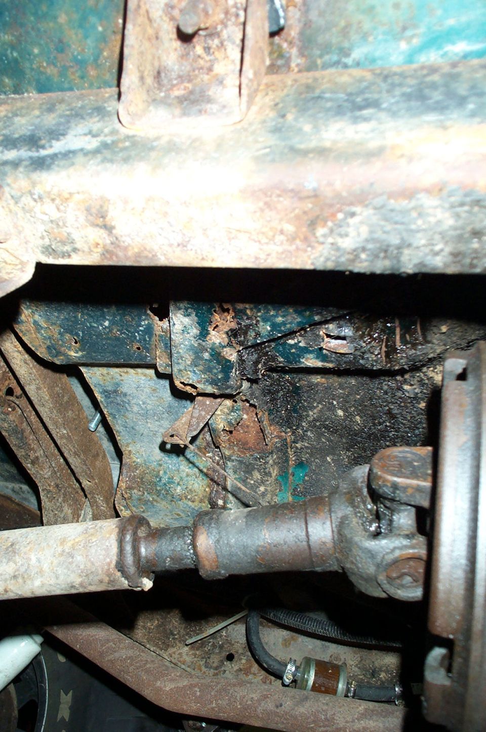 Rusted vehicle underbody with driveshaft, U-joint, and corroded frame showing perforation
