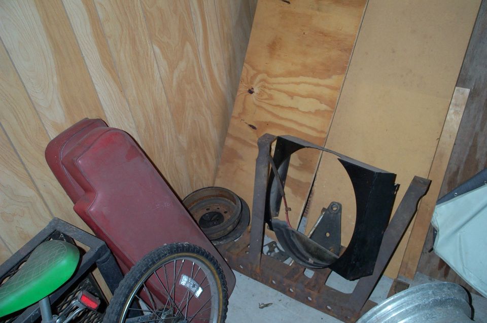 Assorted vehicle parts including red tank, spoked wheel, and black fan shroud in a storage corner