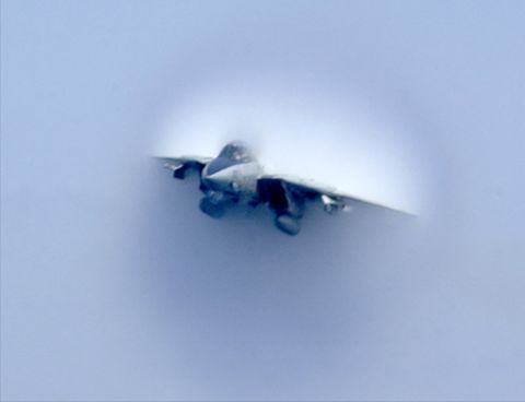 1987 Ford Bronco - A fighter jet breaks the sound barrier, visible through a condensation cone. The Supermotors.net logo is in the lower right corner.