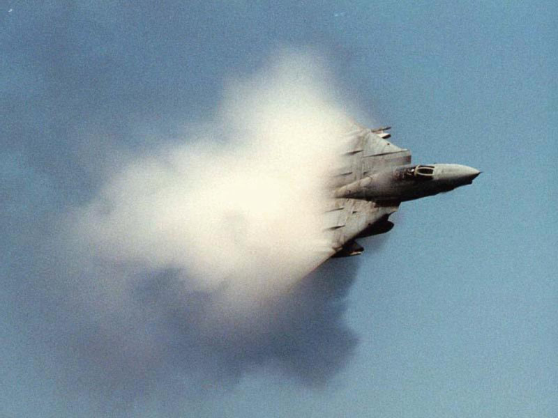 Fighter jet creating a vapor cone as it breaks the sound barrier against a blue sky. Supermotors.net logo visible