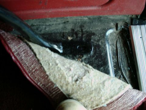 1984 Ford Bronco - The interior floor of a vehicle shows worn carpet and exposed metal, with a dark area suggesting a tire well.