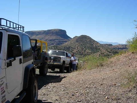 1981 Ford F-150 - A convoy of off-road vehicles navigates a dusty mountain trail under a clear blue sky.
