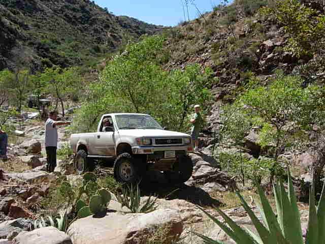 White Toyota pickup truck with lifted suspension driving on a rocky, mountainous trail. Another vehicle is visible in the background