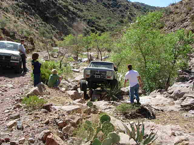 Two off-road vehicles navigating a rocky, dry creek bed with people standing nearby. One vehicle is a Jeep CJ