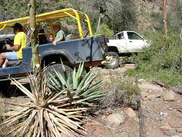 Jeep CJ-style vehicle and white pickup truck on a rocky, overgrown trail