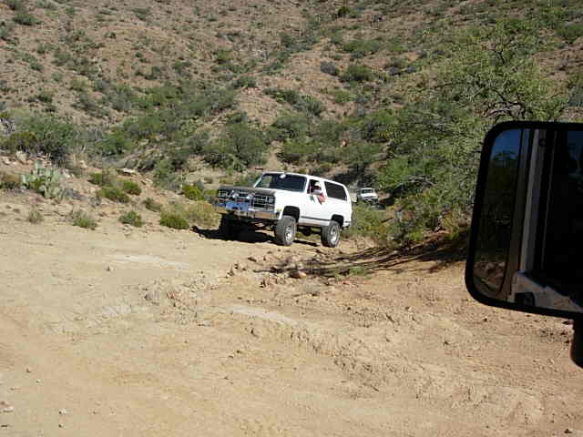 White SUV with chrome grille and bumper on a dirt road, side view. Another white vehicle is visible in the distance