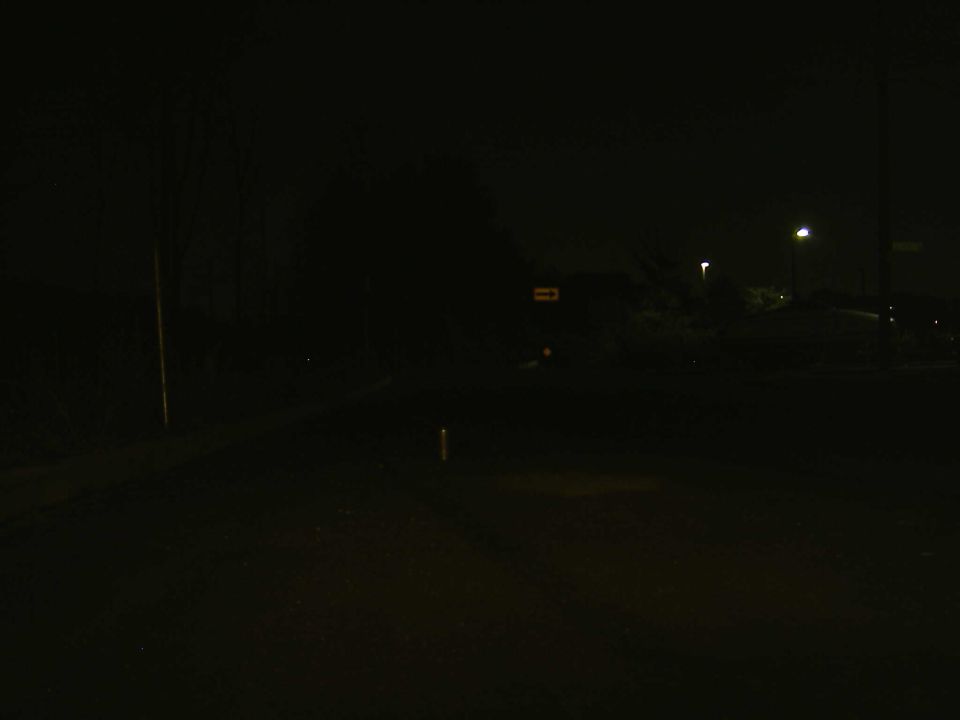 Very dark night scene of a road with distant streetlights and a faint arrow sign; no vehicle visible