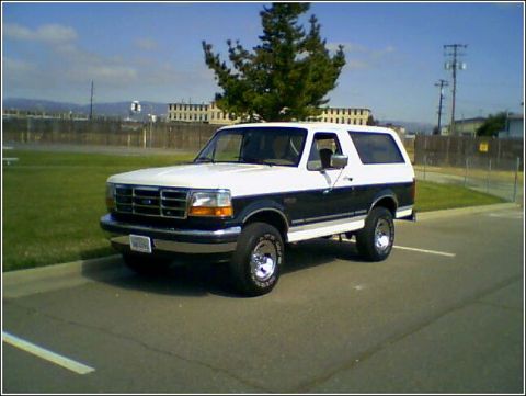 1995 Ford Bronco - A two-tone Ford Bronco is parked in an asphalt lot with a grassy area and fence in the background.