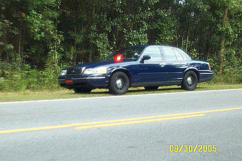 1998 Ford Crown Victoria - Dark blue Ford Crown Victoria sedan parked on the grass shoulder of a two-lane road with a red beacon light glowing on the roof and a red l…