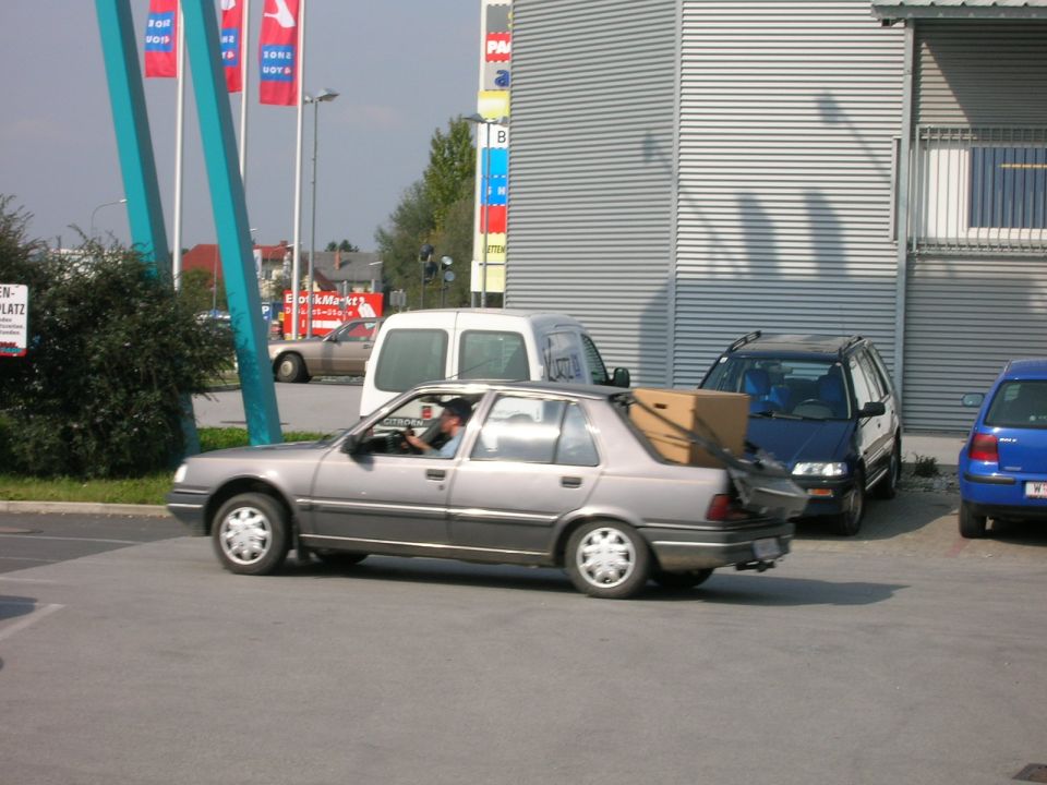 Silver hatchback driving in a store lot with a large cardboard box strapped in the open hatch