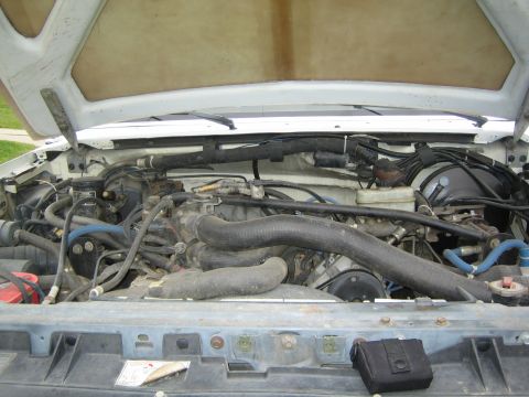 1990 Ford Bronco - Full engine bay view under an open hood showing the radiator hose, brake master cylinder, wiring and a black battery near the core support.