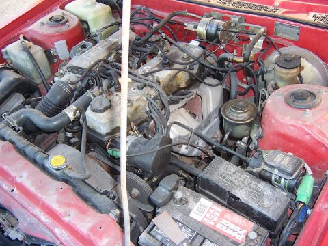 1984 Toyota Celica - The engine bay of a red 1984 Toyota Celica shows a V6 engine, battery, radiator, and various hoses and wires.